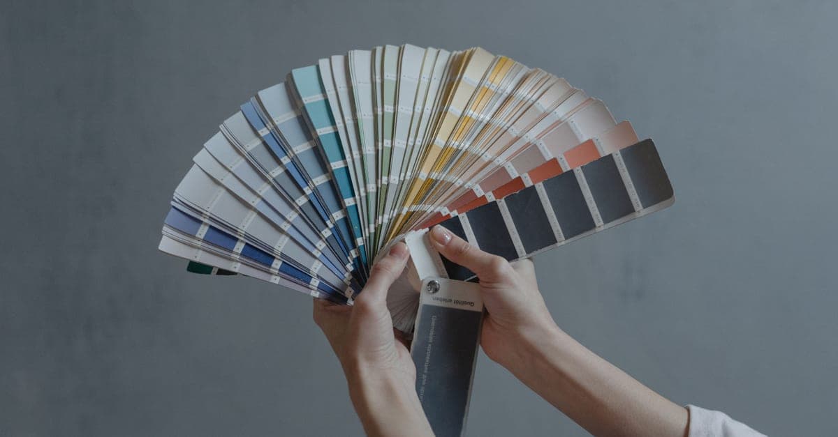 Hands holding an open fan of colour swatches against a plain grey background.