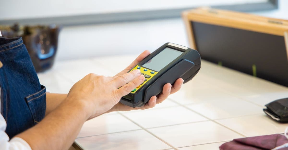 A shop assistant in an apron holding a card payment terminal above a tiled counter.