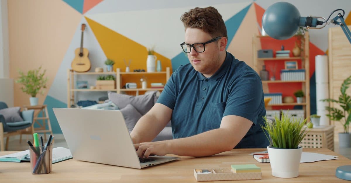 A man working at a laptop in a bright room with a boldly painted geometric wall behind him.