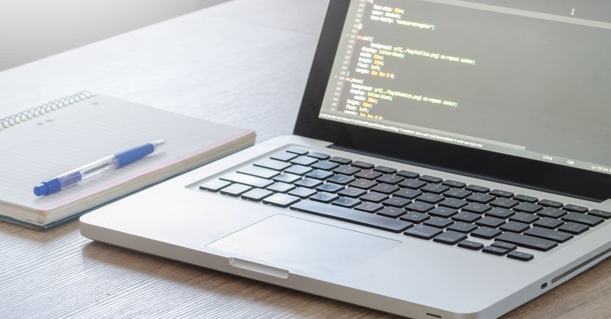 A laptop open on a code editor beside a notebook and pen on a wooden desk.