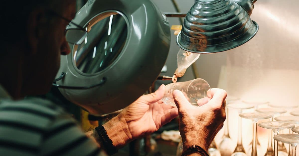 A craftsman inspecting a glass under a large magnifying lamp, with rows of finished glasses beside him.