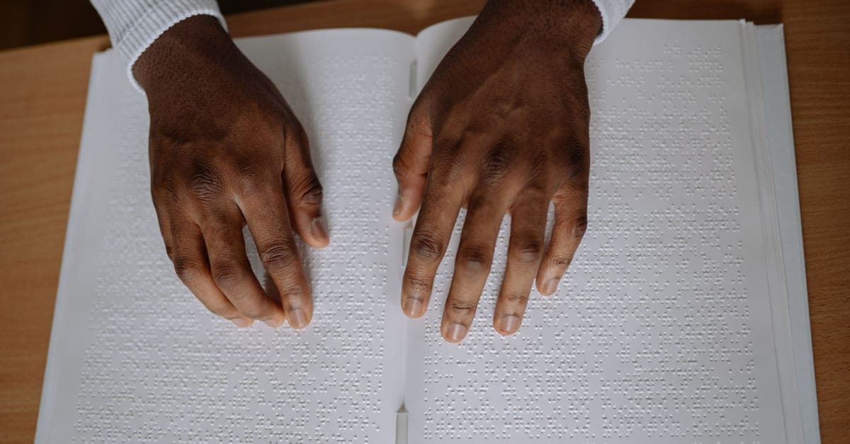 A pair of hands reading a page of braille in an open book on a wooden table.
