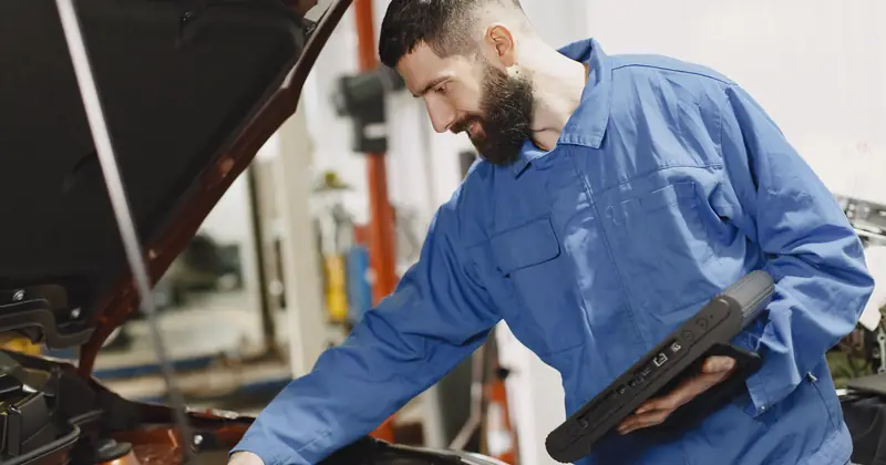 An auto mechanic in blue coveralls inspecting a car's engine in a garage.