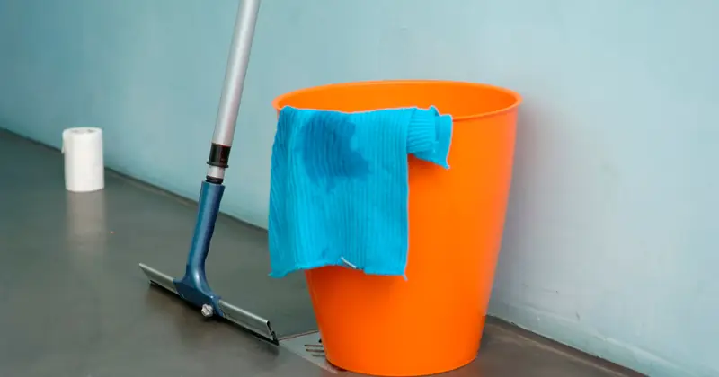 Close-up of cleaning supplies including orange bucket, mop, and blue rag indoors.