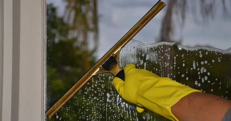 Close-up of hand cleaning window with squeegee outdoors using yellow gloves.