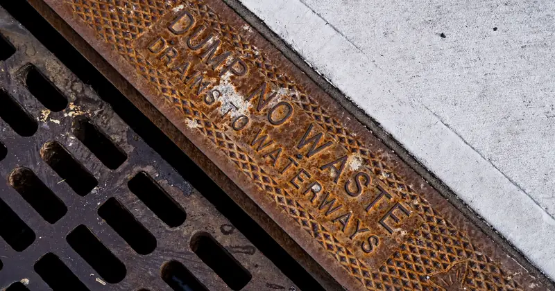 Close-up of a rusty drainage grate with environmental warning text on concrete sidewalk