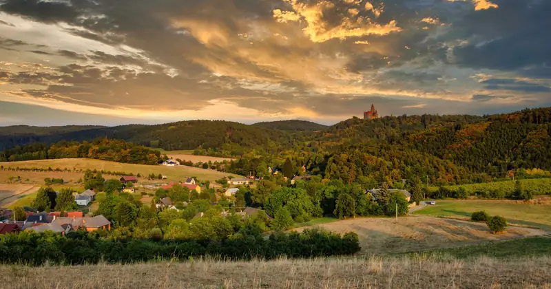Scenic view of a tranquil Czech village surrounded by lush hills at sunset, under an orange sky.