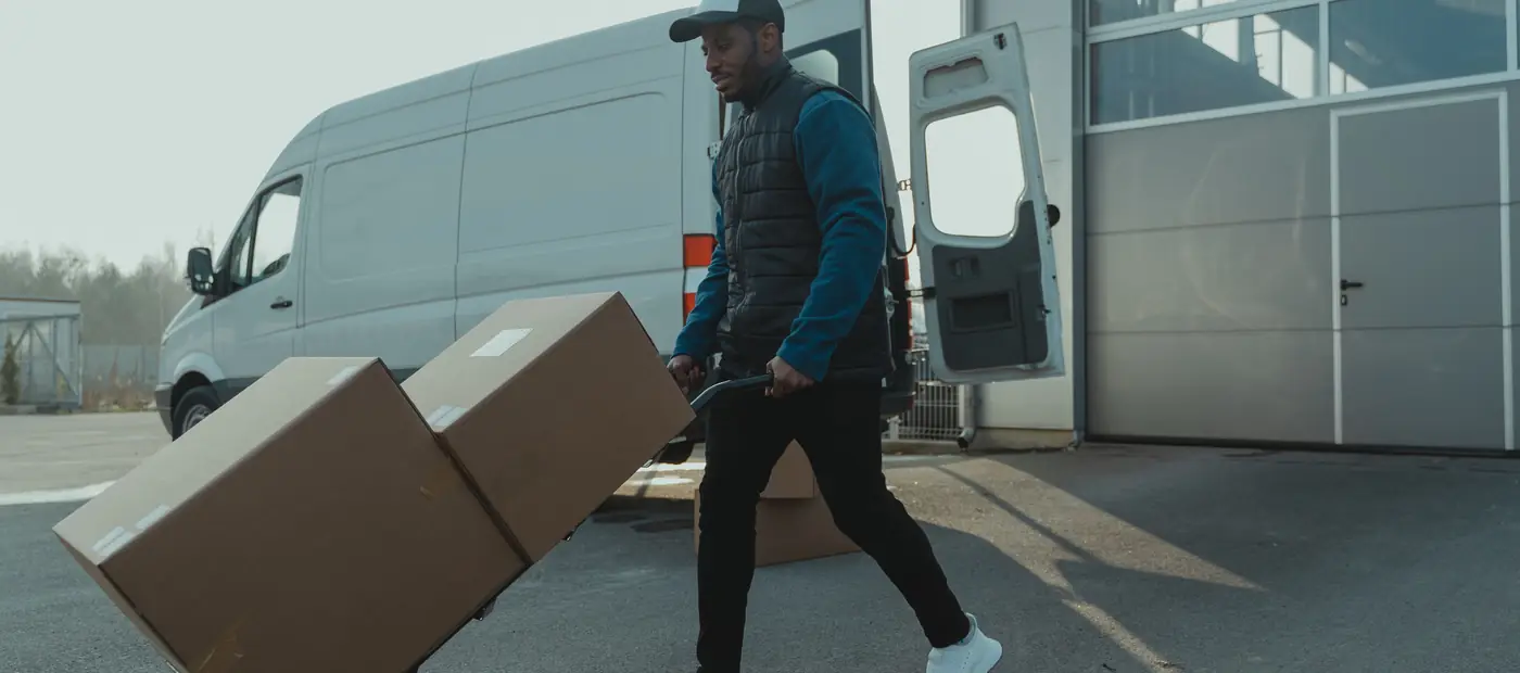 Courier pushing a trolley with boxes near a van at a warehouse, showcasing delivery services.