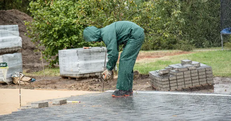 A worker in protective gear lays paving stones on a wet day.