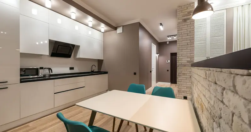 Table with chairs placed in modern kitchen with white cupboards and counter with appliances in spacious apartment with glowing lights