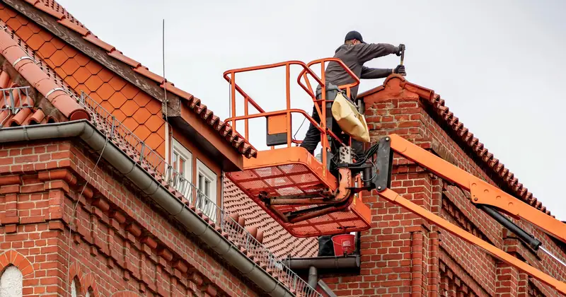 Roofer in a cherry picker conducting maintenance on a red brick building with a tiled roof.