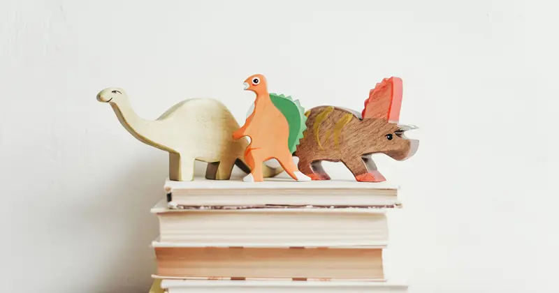 Colorful wooden dinosaurs on top of a stack of hardcover books on a light background.